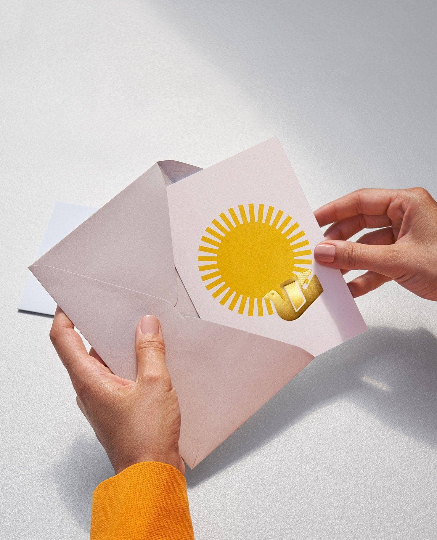 A close-up of hands holding an open pale pink envelope or card, with a yellow sun illustration and a small gold ribbon detail visible on the front, against a light gray background.