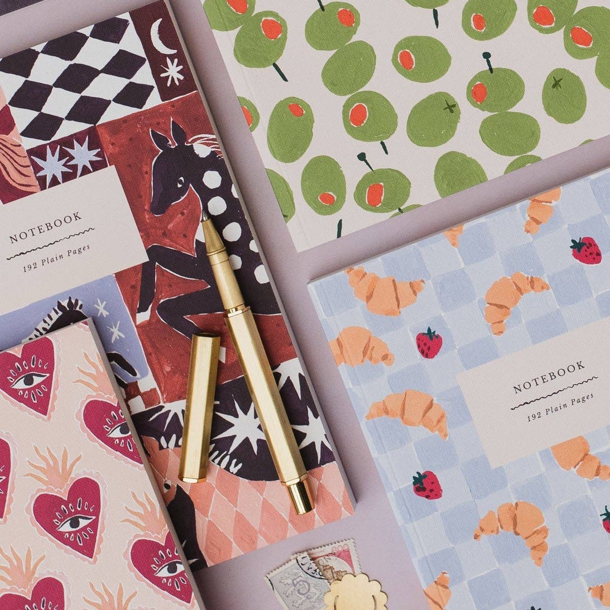 A styled flat-lay of multiple patterned notebooks with a gold pen placed across them on a pale lavender background. The notebooks feature colorful designs including green olives, pink hearts, orange croissants, black-and-white check, and a dark horse illustration.