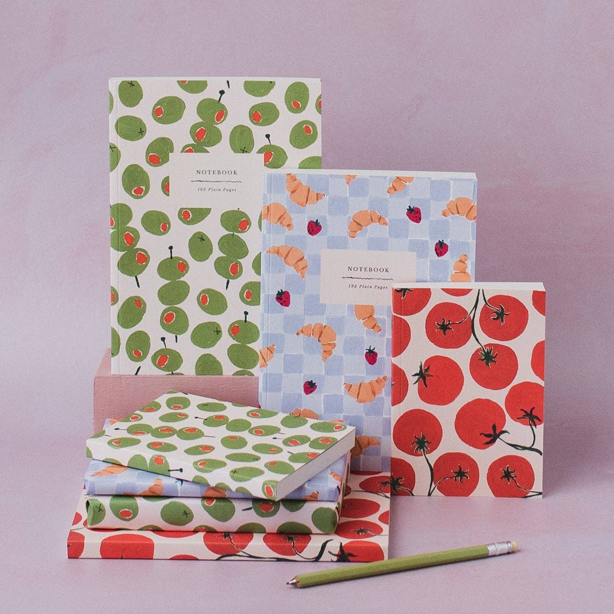 A set of patterned notebooks and wrapping paper displayed on a dusty pink background, featuring green avocado prints, orange and red strawberry checks, and red tomatoes. A green pencil rests at the bottom.
