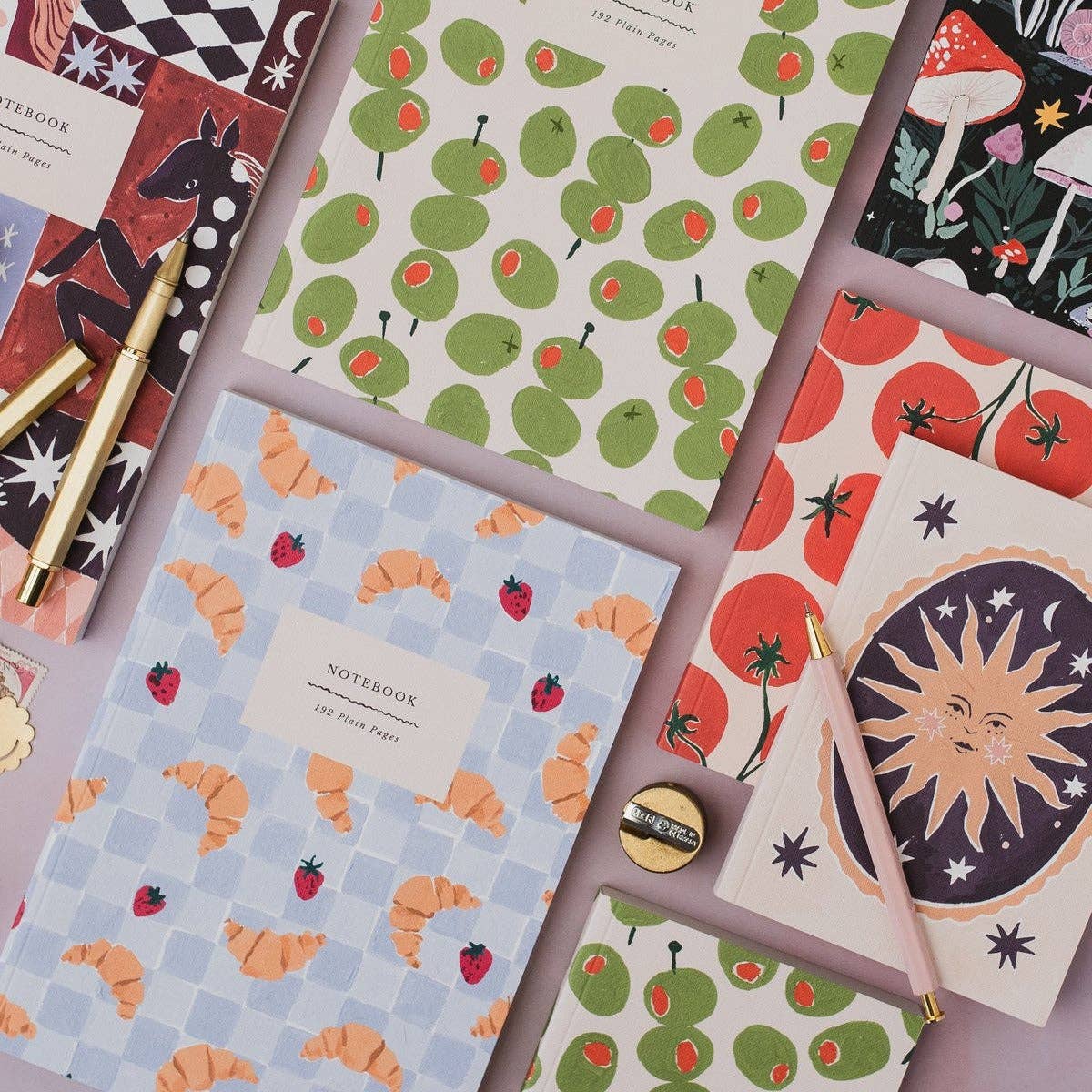 A styled flat-lay of multiple patterned notebooks and stationery items arranged on a pale pink background. Visible items include notebooks with green apple prints, red strawberry motifs, orange crescent moon designs, a mushroom forest print, a sun illustration, gold and pink pens, and a small gold pencil sharpener.