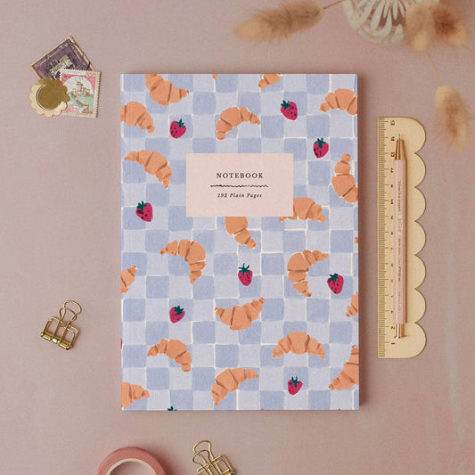 A notebook with a pastel blue and lavender checkered pattern, decorated with orange croissants and red strawberries, displayed in a styled flat-lay with a yellow scalloped ruler, gold binder clips, and decorative paper on a dusty pink background.