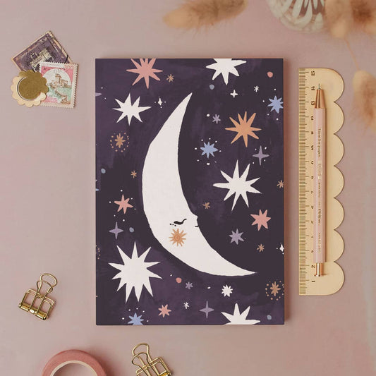 Notebook with celestial design featuring a moon and stars on a pink surface with stationery items.