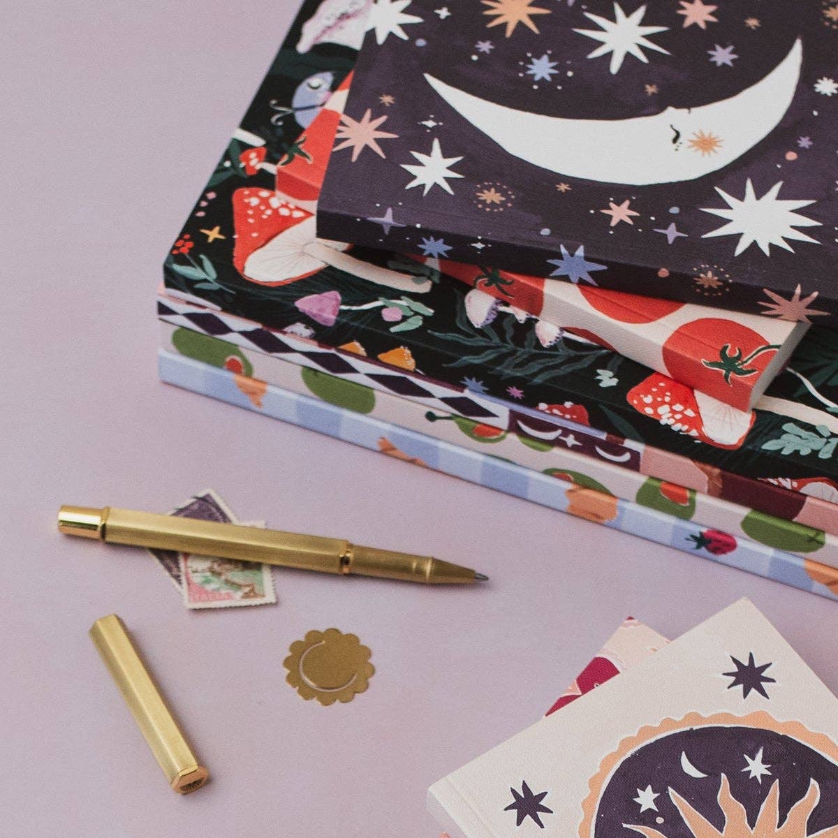 A stack of decorative notebooks with whimsical illustrated covers, including a crescent moon, stars, mushrooms, and floral patterns, arranged on a pale pink surface with a gold pen and small stationery pieces nearby.