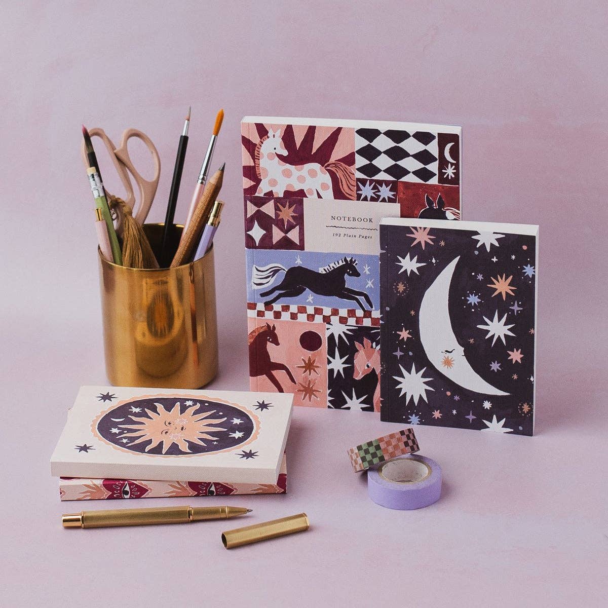 A styled flat-lay of stationery items on a soft pink background, including patterned notebooks, a gold pencil cup with pens and scissors, washi tape, and gold pen parts.