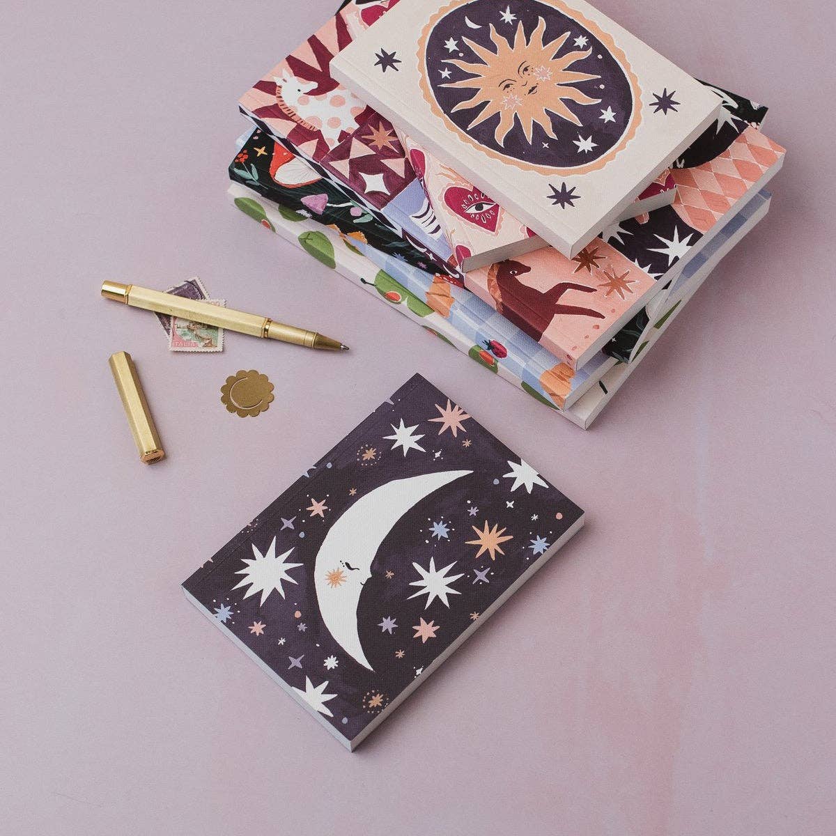 A stack of patterned notebooks and a separate moon-and-stars notebook are arranged on a soft pink background with a gold pen, small card, and gold seal nearby.