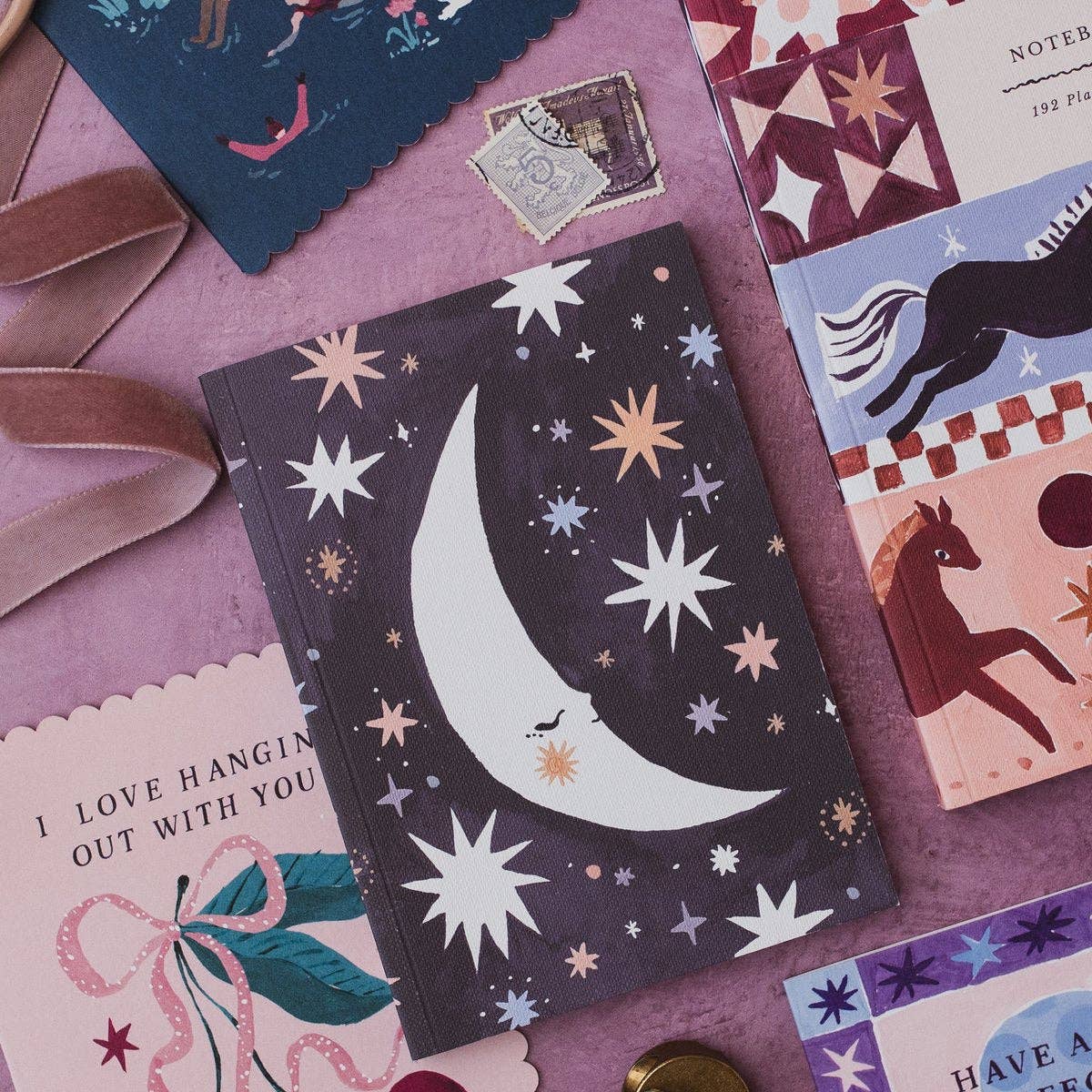 A decorative notebook with a dark purple cover featuring a large white crescent moon and scattered stars, styled on a pink surface with other stationery items around it.