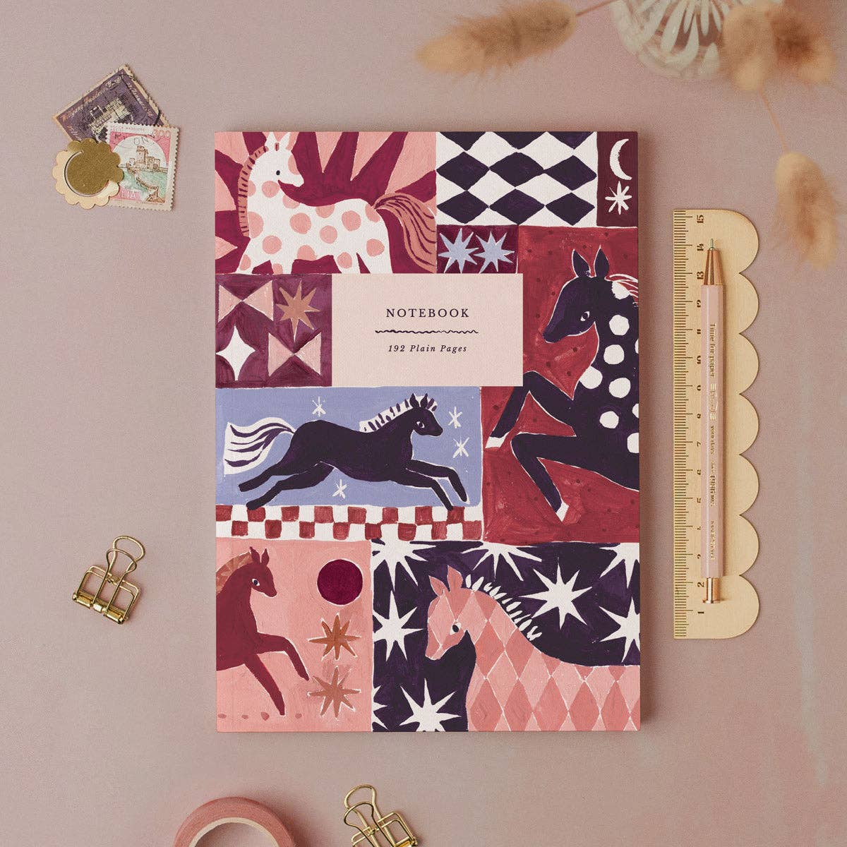 A patterned notebook with horse illustrations lies on a dusty pink surface, styled with a scalloped ruler, binder clips, paper clips, and small decorative items.