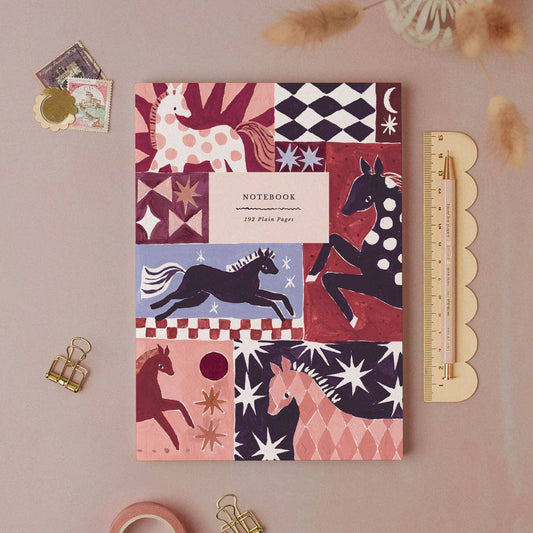 A patterned notebook with horse illustrations lies on a dusty pink surface, styled with a scalloped ruler, binder clips, paper clips, and small decorative items.