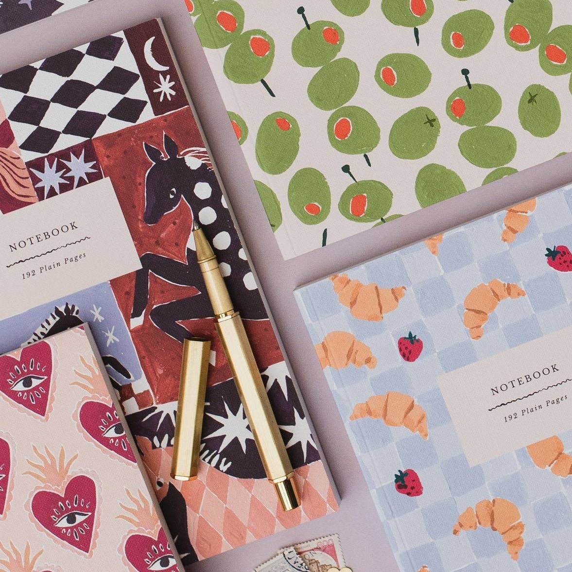 A styled flat-lay of four patterned notebooks with a gold pen placed across them on a pale lavender background. The notebooks feature whimsical illustrations including green olives, pink hearts, strawberries, croissants, a horse, and a black-and-white check pattern.