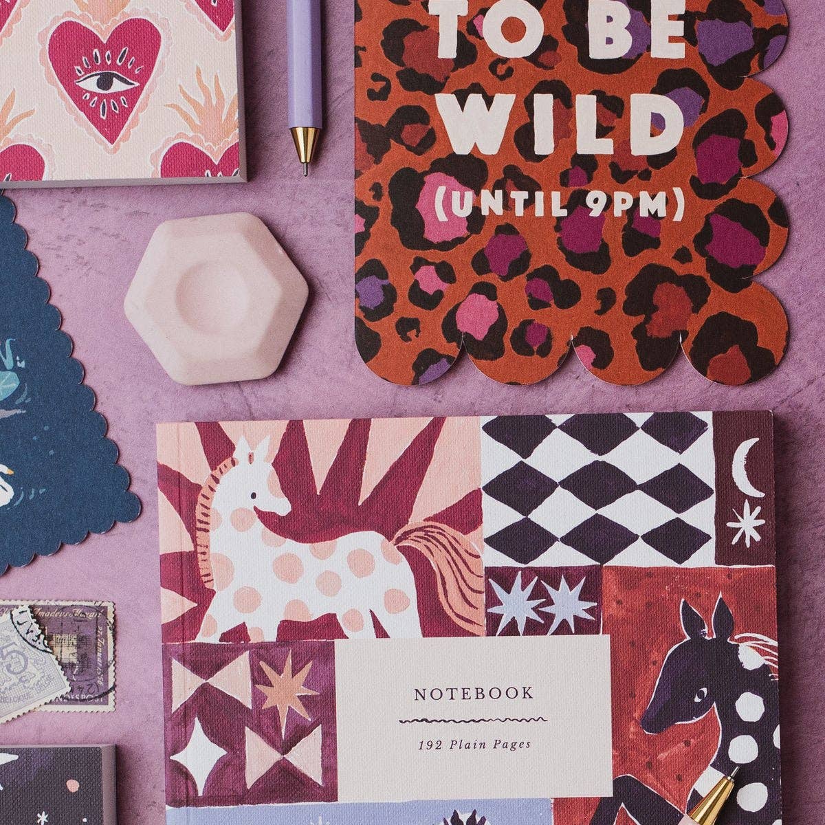 A styled flat-lay of stationery items on a textured lavender surface, including a patterned notebook with a unicorn and geometric blocks, a pink heart notebook, a purple pen, a small pale pink hexagonal object, and a scalloped sign reading 'BORN TO BE WILD (UNTIL 9PM)'.
