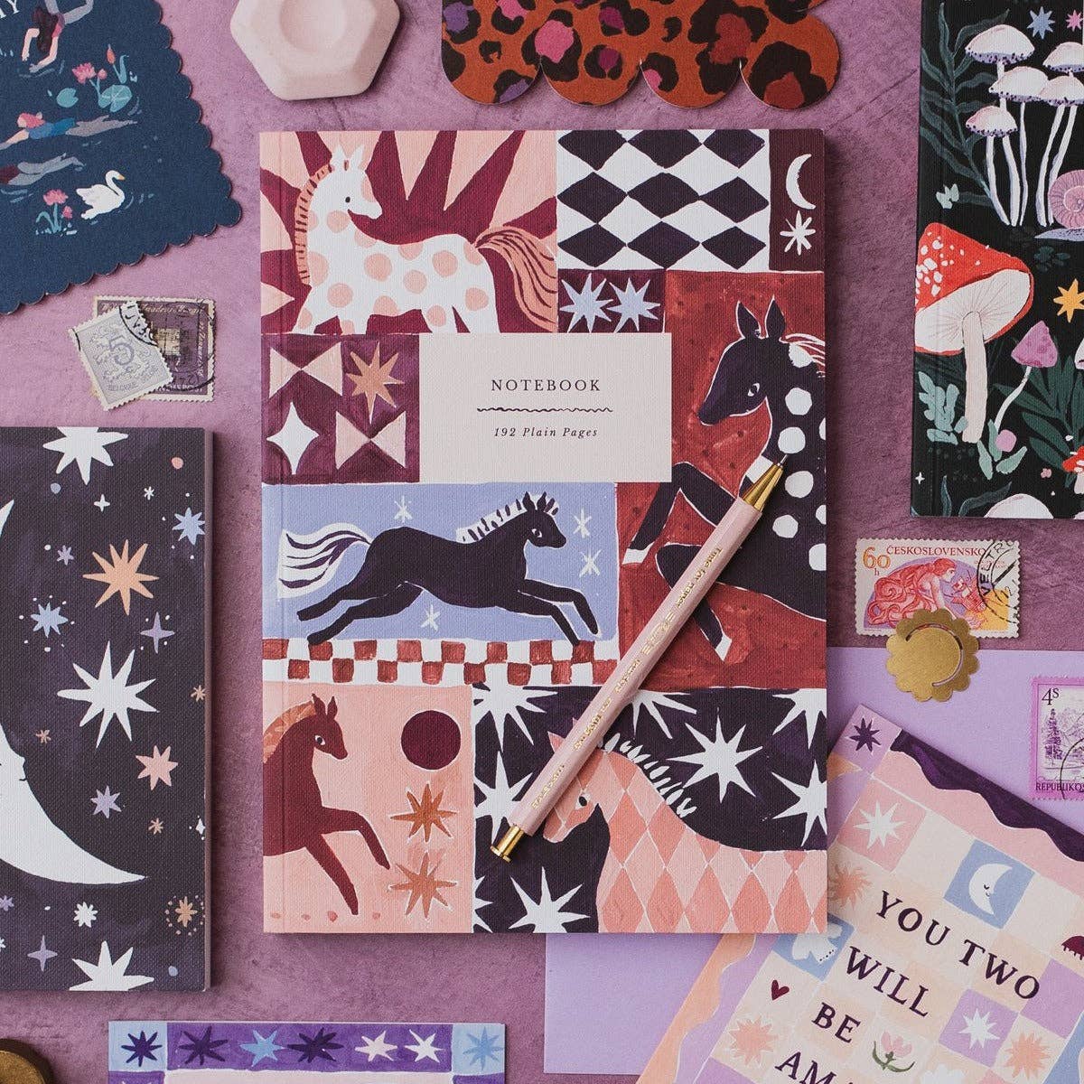 A patchwork-patterned notebook with illustrations of horses, stars, moons, and mushrooms, styled in a flat lay with a gold pen on a purple surface.
