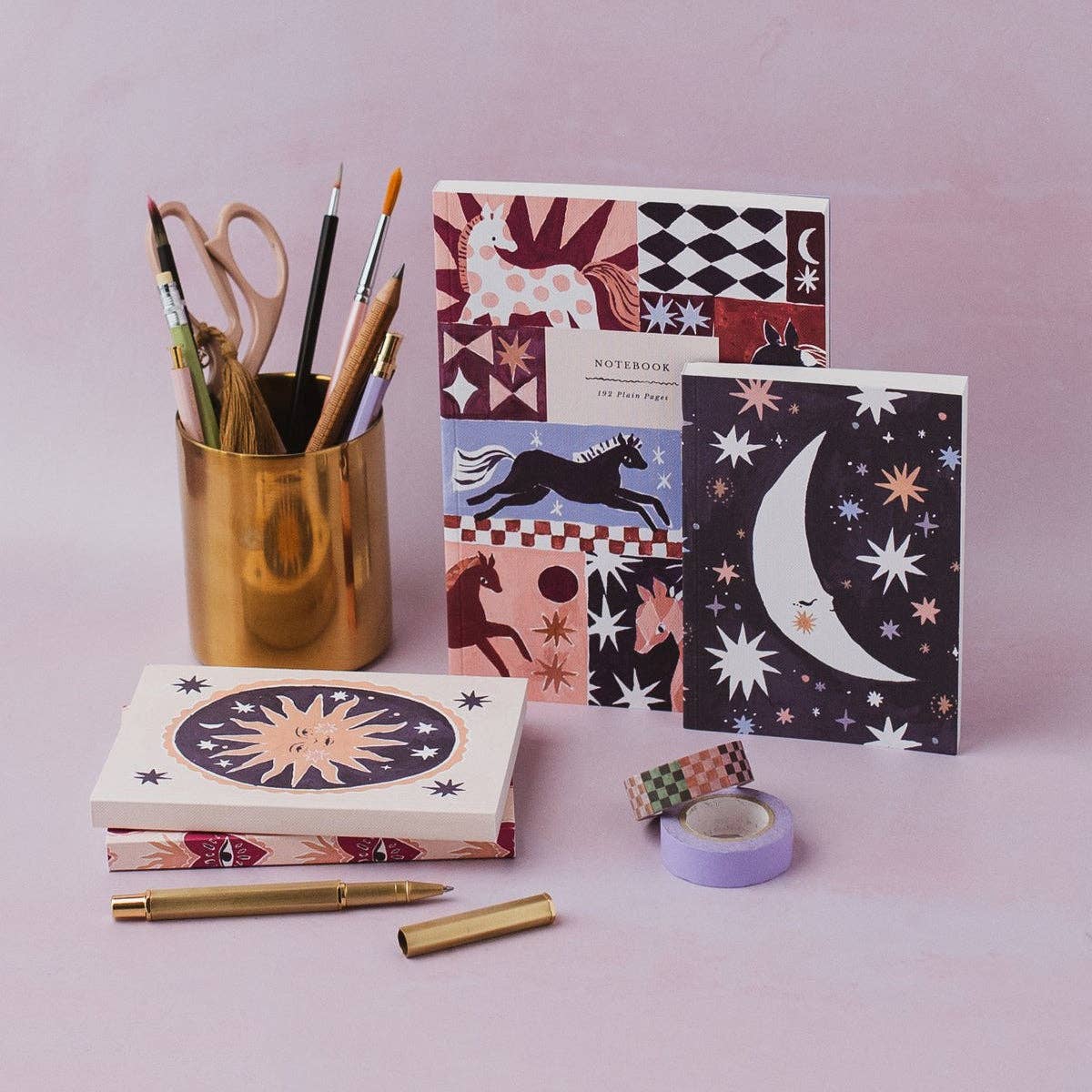A styled flat-lay of stationery items on a soft pink background, including a gold pen, gold pencil, a gold pencil cup with paintbrushes and scissors, a notebook, a patterned notebook, washi tape rolls, and a small decorative item.
