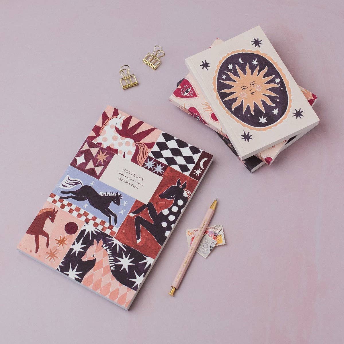 A styled flat-lay of a notebook with a multicolor patchwork horse pattern, a matching decorative box with a sun illustration, a pink pen, and small gold binder clips on a soft pink background.
