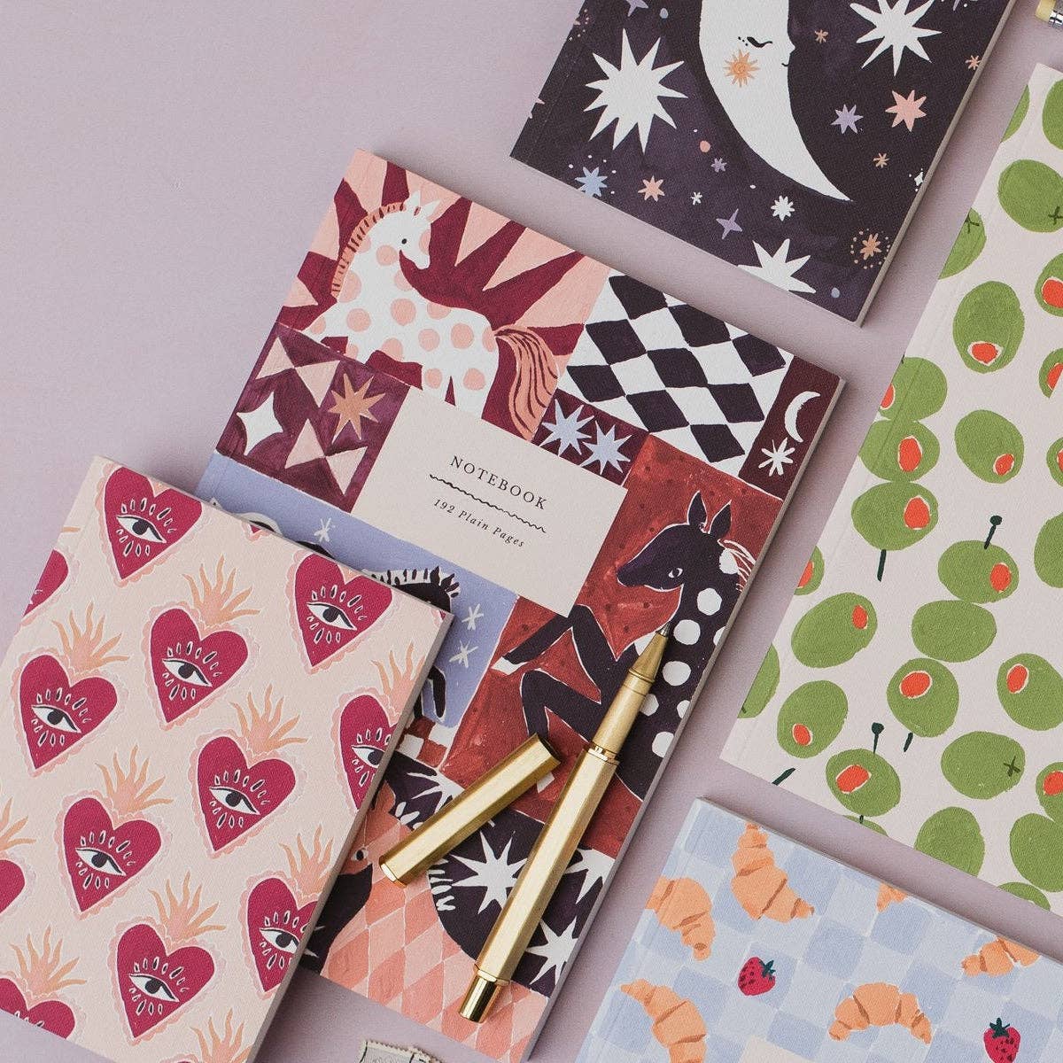A styled flat-lay of several patterned notebooks with a gold pen on top, arranged on a light lavender background. The notebooks feature whimsical designs including hearts with eyes, a unicorn, stars and moons, olives, and croissants.