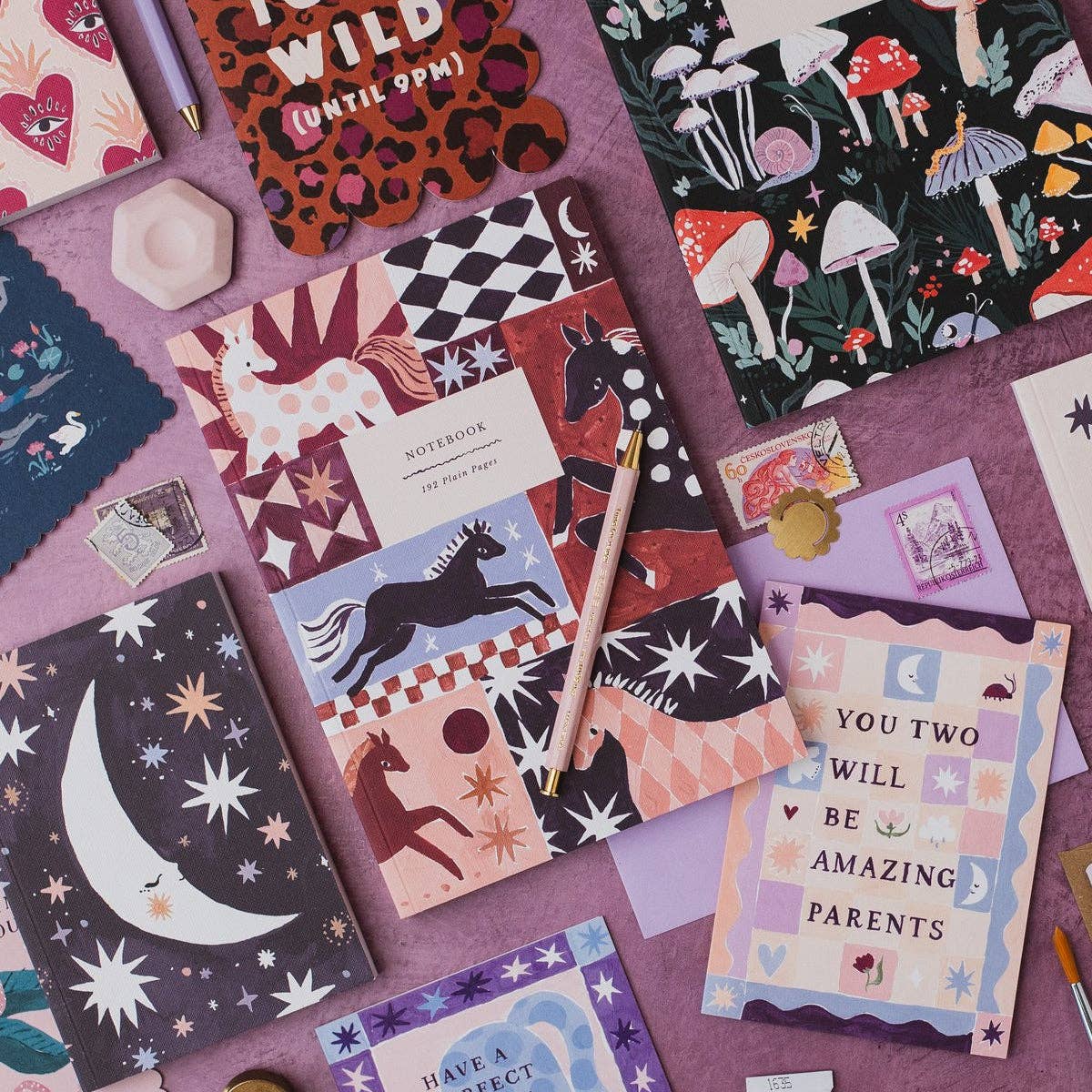 A styled flat-lay of multiple patterned notebooks and a matching card set arranged on a purple surface. The products feature whimsical illustrations including unicorns, horses, mushrooms, stars, moons, and text such as 'WILD (UNTIL 9PM)' and 'YOU TWO WILL BE AMAZING PARENTS'.