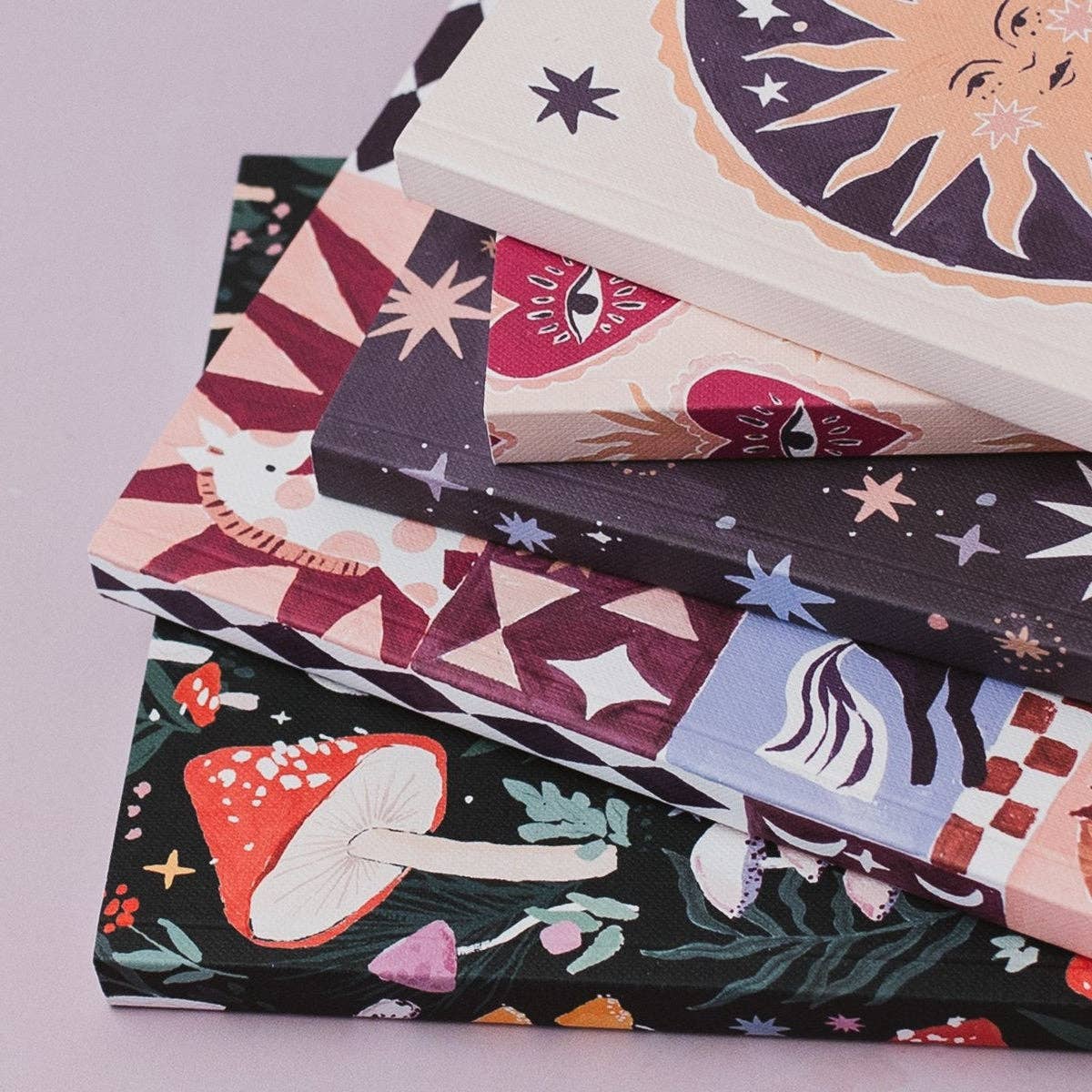 A stack of decorative notebooks or journals with whimsical illustrated covers, including stars, a sun, an eye motif, a white cat, mushrooms, and abstract patterns, photographed on a soft light lavender background.