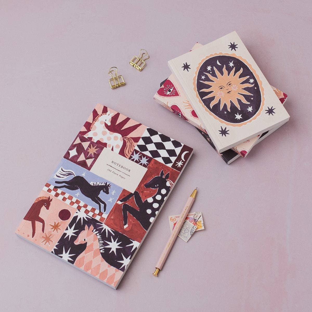 A styled flat-lay of two patterned notebooks on a dusty pink background, accompanied by gold binder clips, a wooden pen, and a small star-shaped paper weight. One notebook features a colorful patchwork of horses, stars, and geometric designs, while the other displays a large sun illustration on the cover.