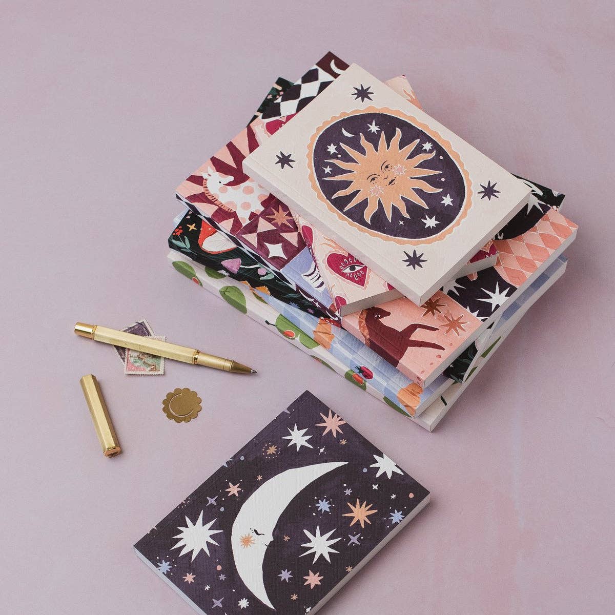 A stack of patterned notebooks or cards with whimsical celestial and animal illustrations, arranged on a soft pink background alongside a gold pen, a small gold seal, and another notebook featuring a crescent moon and stars.