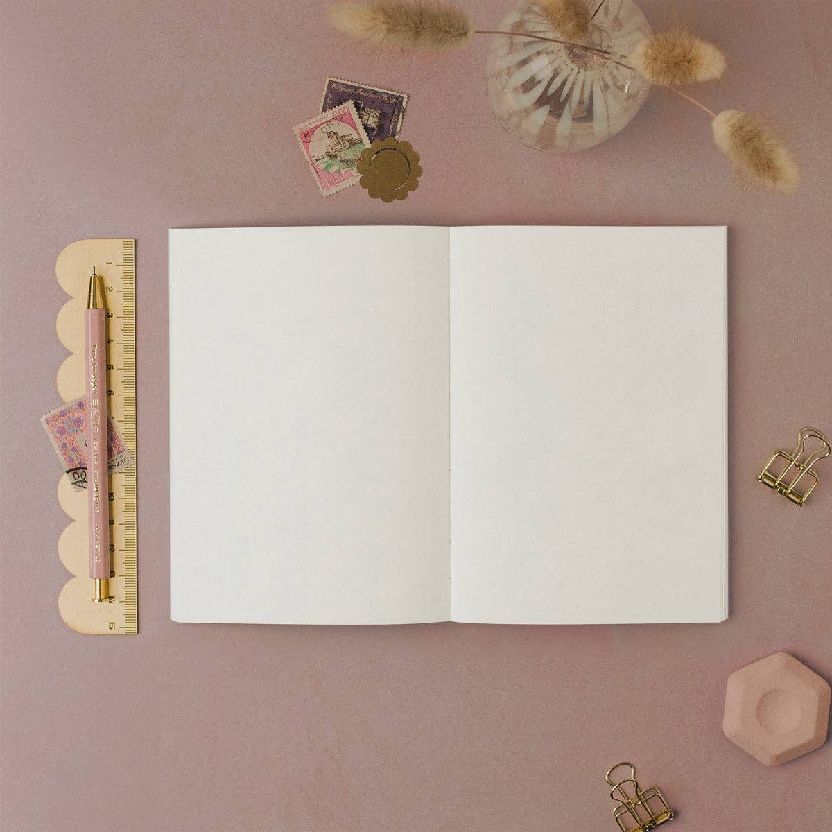 An open blank notebook with white pages lies on a dusty pink background, styled with stationery props including a pink pen, a scalloped ruler, small stamps, clips, and dried grass.