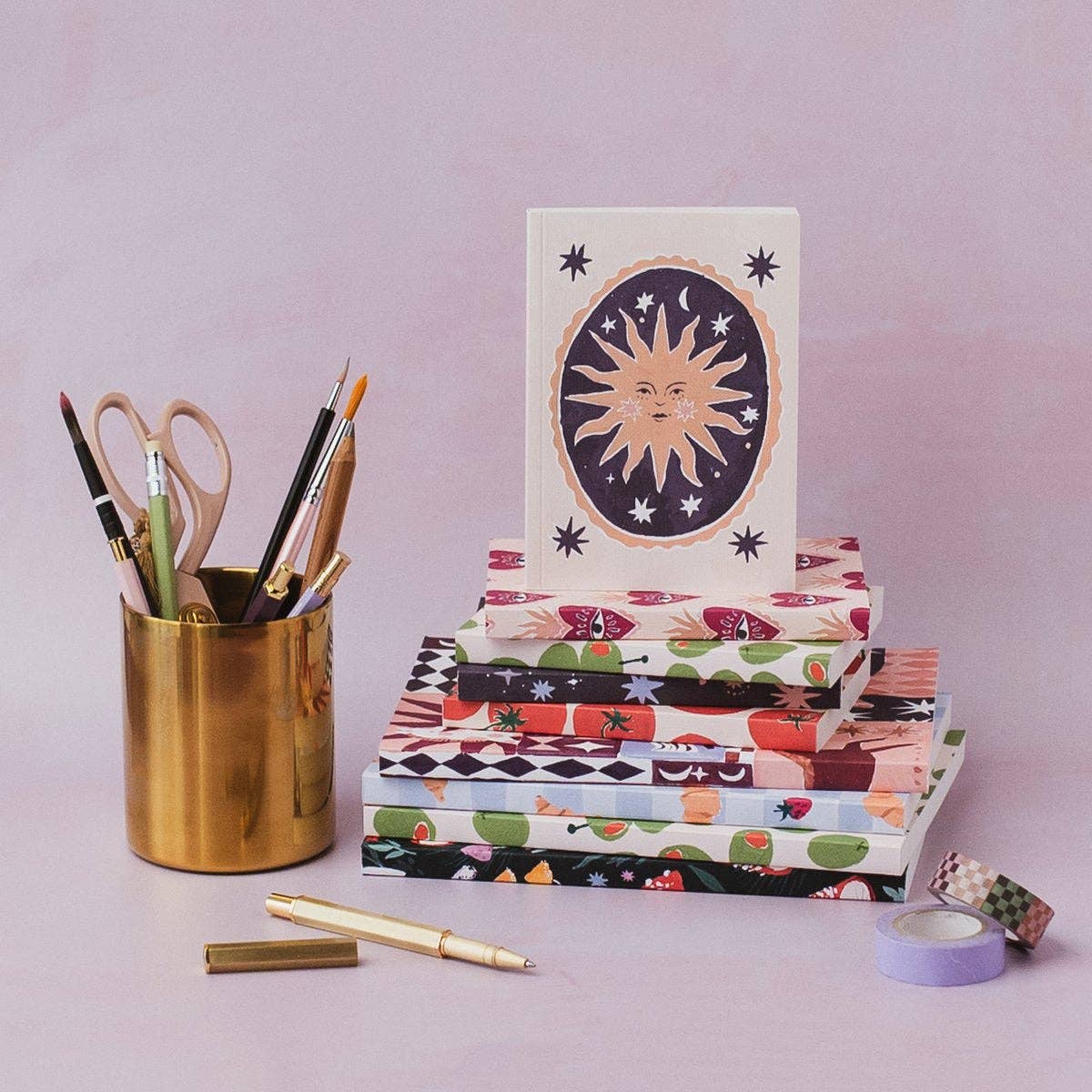 A stack of patterned paper notebooks or cards with a sun illustration standing on top, arranged on a pale pink surface next to a gold pen, a gold pencil holder with pens and scissors, and washi tape.