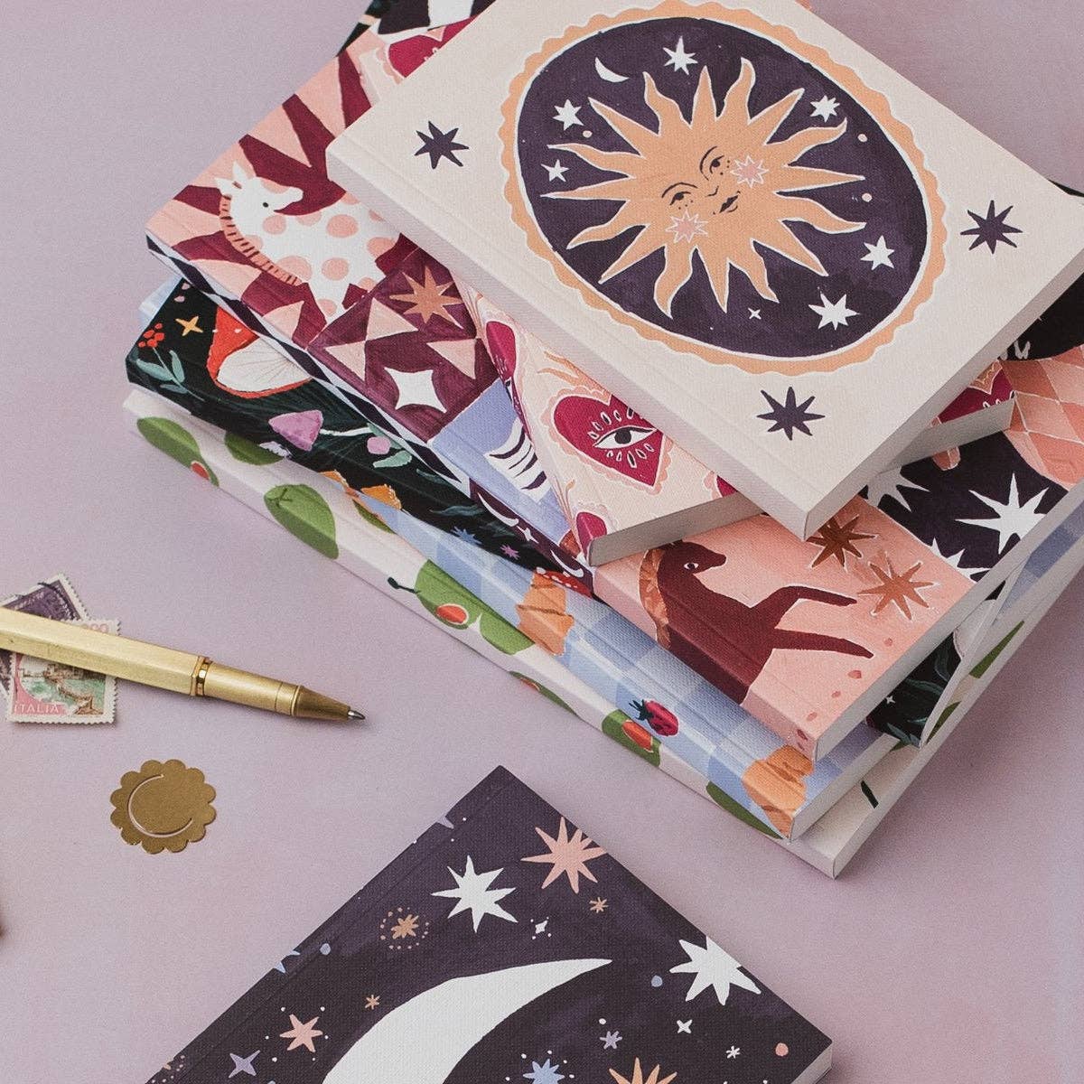 A stack of patterned notebooks or journals with whimsical illustrations, including a sun, stars, animals, and abstract shapes, arranged on a soft pink surface with a gold pen and small decorative items nearby.