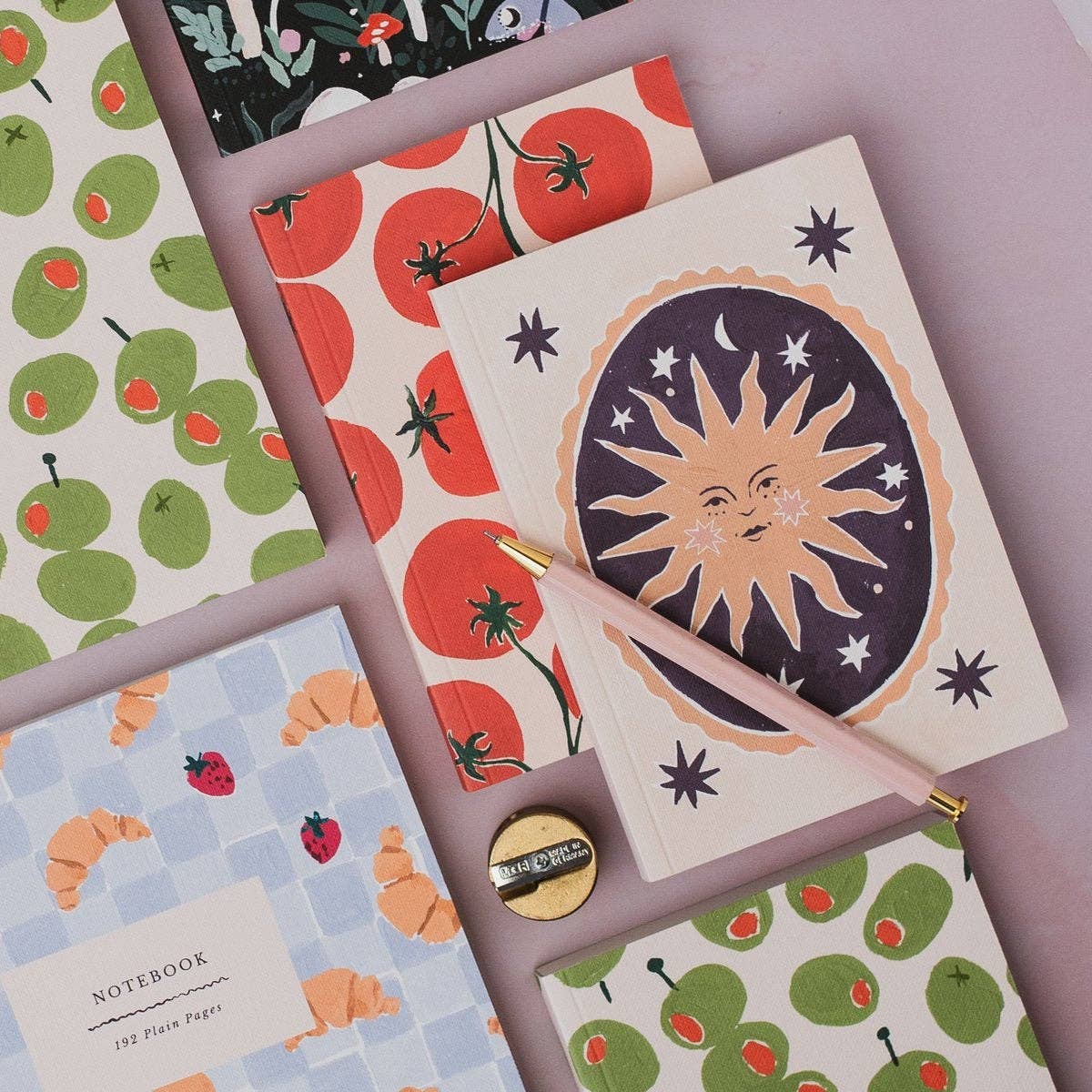 A styled flat-lay of patterned notebooks and a pink pen on a soft lavender background. The notebooks feature whimsical illustrations including a smiling sun with stars on one cover, red tomatoes on orange stems on another, green apples with red details on a third, and croissants on a light blue checkered pattern on a fourth.
