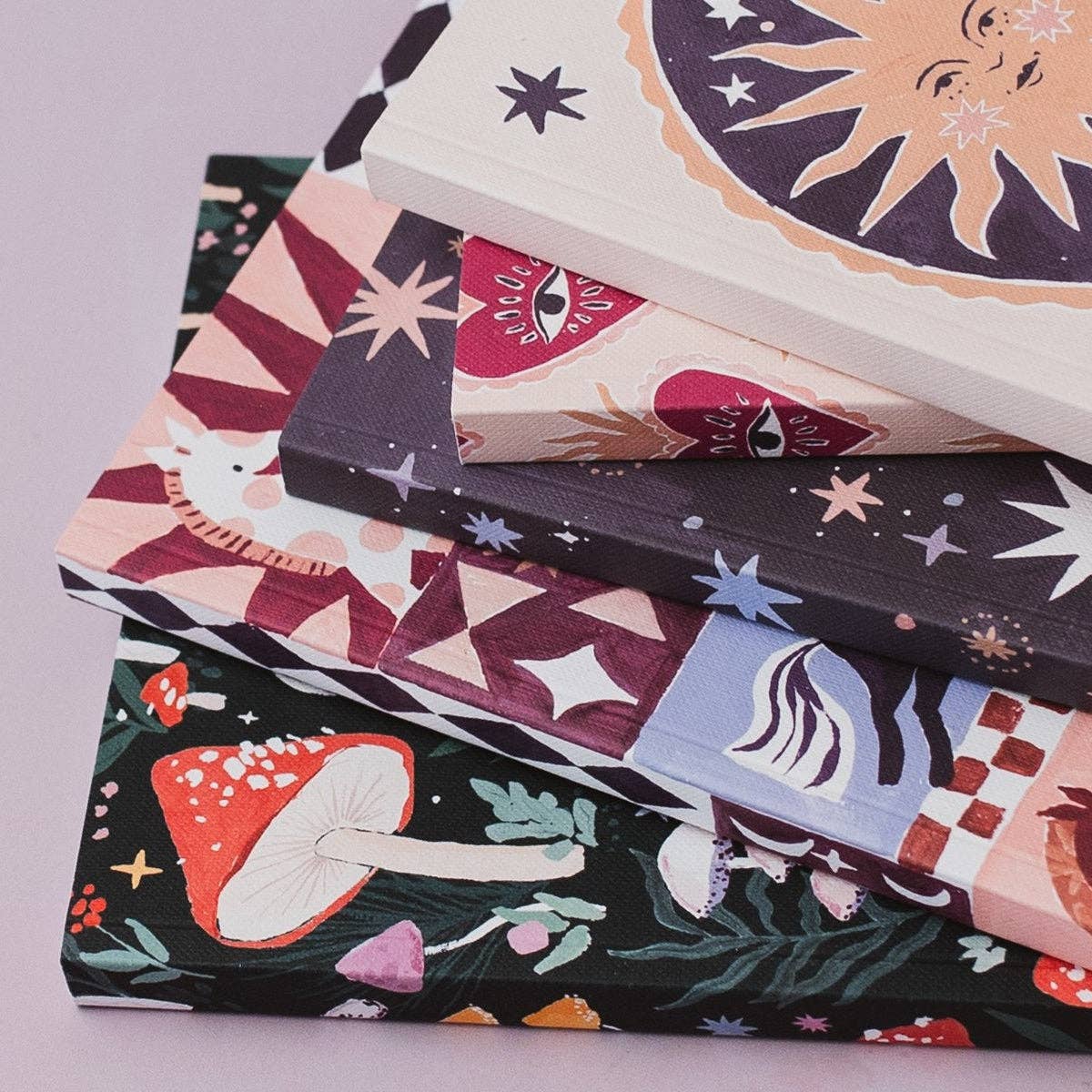 A stack of patterned notebooks or book covers with whimsical illustrations, including a sun, eye motifs, mushrooms, stars, and geometric shapes, photographed on a soft lavender background.