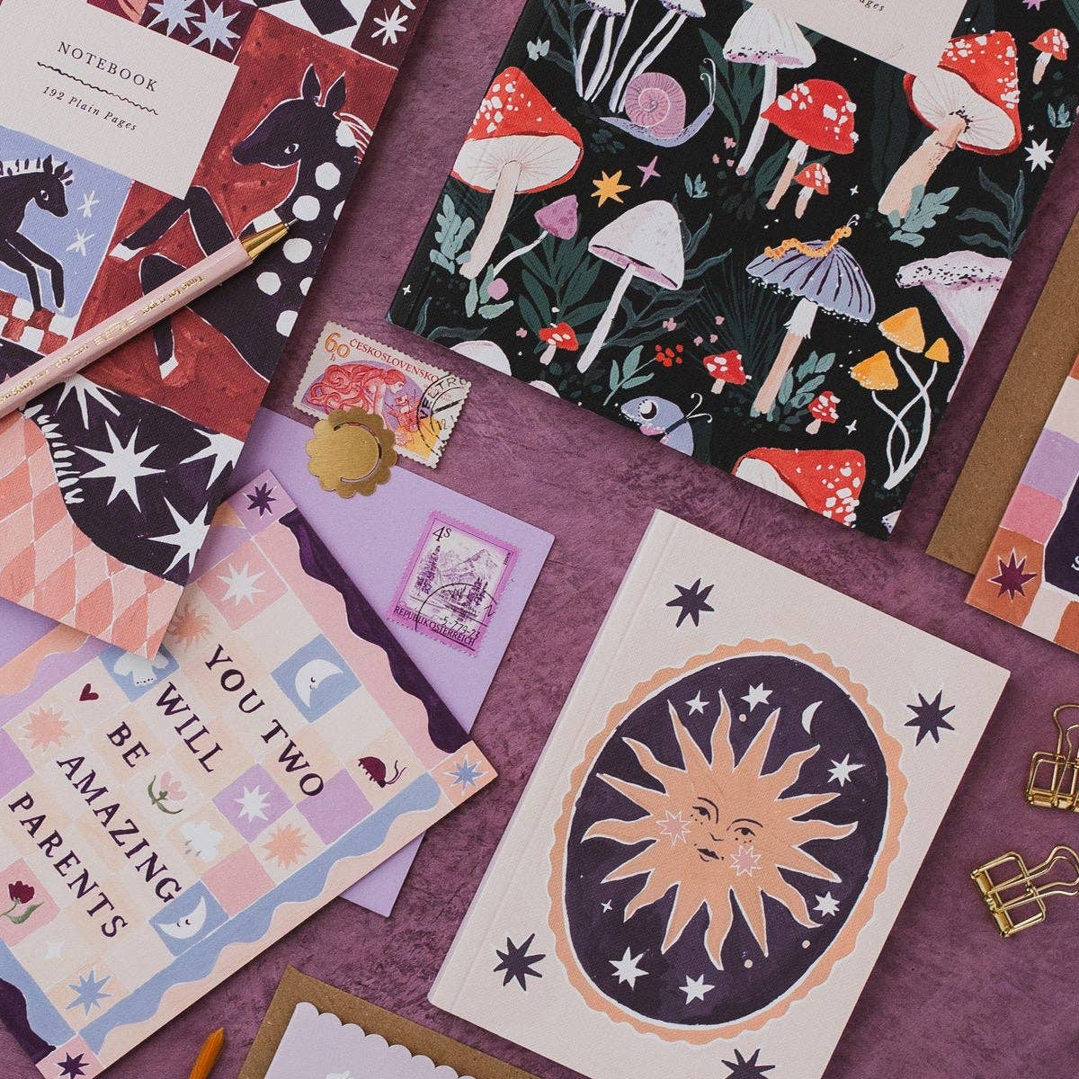 A styled flat-lay of various stationery items, including patterned notebooks, a card with a smiling sun and moon, a purple envelope, and a gold pen, arranged on a textured purple surface.