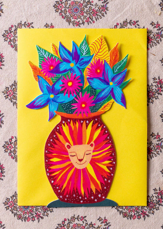 A colorful paper-cut style art print or card featuring a yellow background with a stylized flower vase. The vase is red and yellow with a smiling face, and it holds bright blue, pink, purple, green, and orange flowers and leaves.