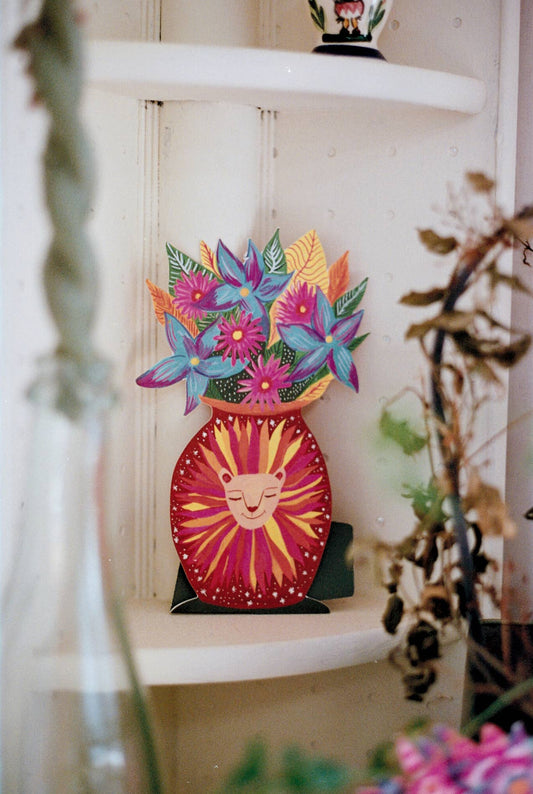 A colorful decorative wooden wall hanging shaped like a sunburst vase filled with stylized flowers, displayed on a white shelf in a softly blurred interior setting.