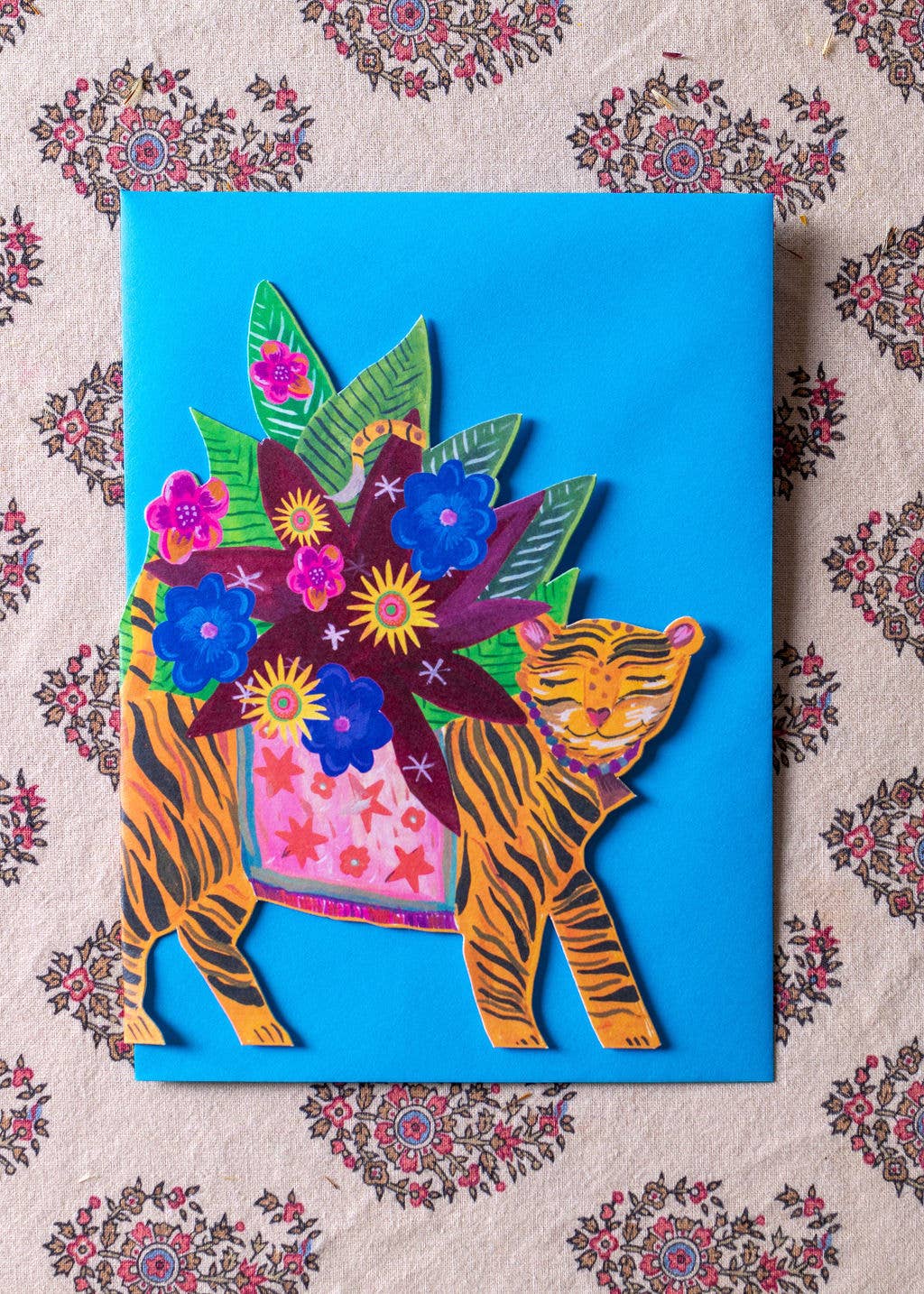 Colorful paper craft of a tiger with flowers on a blue background, placed on a patterned fabric.