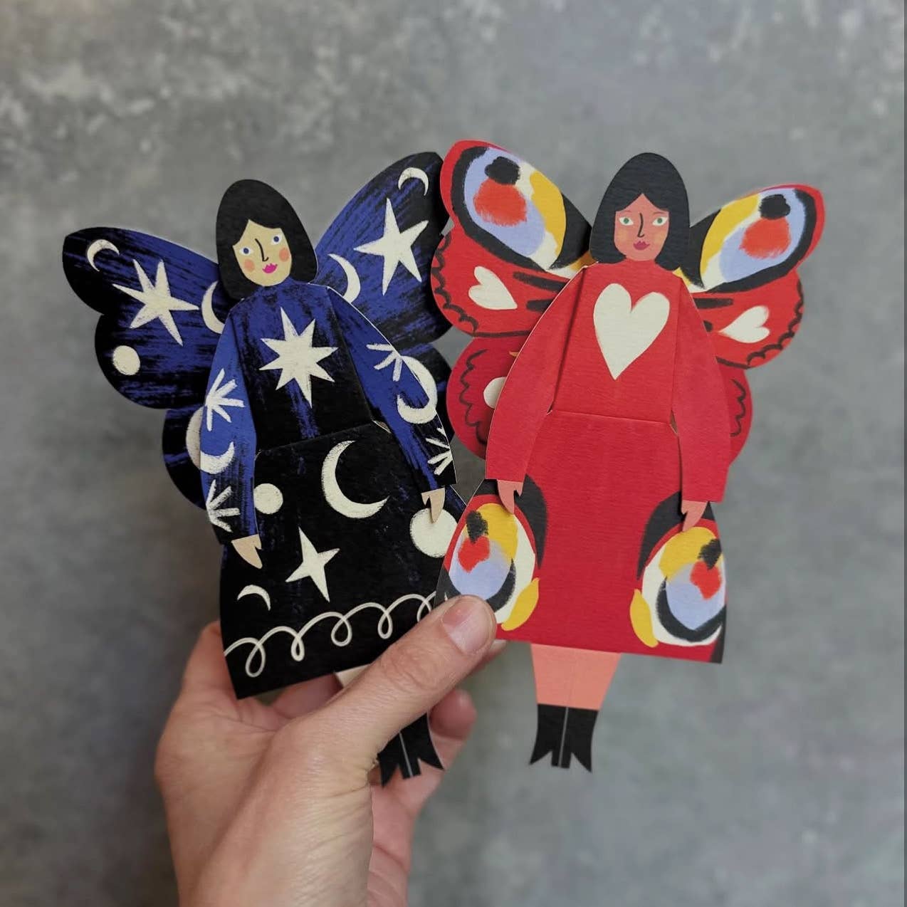Hand holding two colorful paper figures with butterfly wings against a gray background